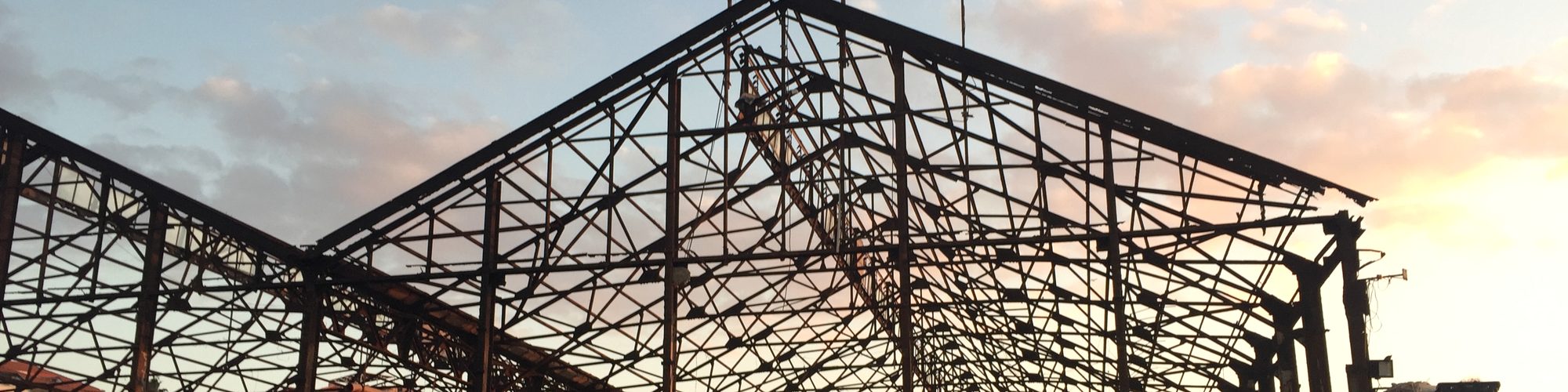 A bare iron roof frame against an evening sky: the structure before the walls